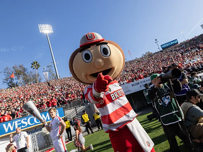 Ohio State's Brutus Buckeye mascot points at the camera from the football field on a bright, blue-skies kind of day. Fans fill the stands behind him.