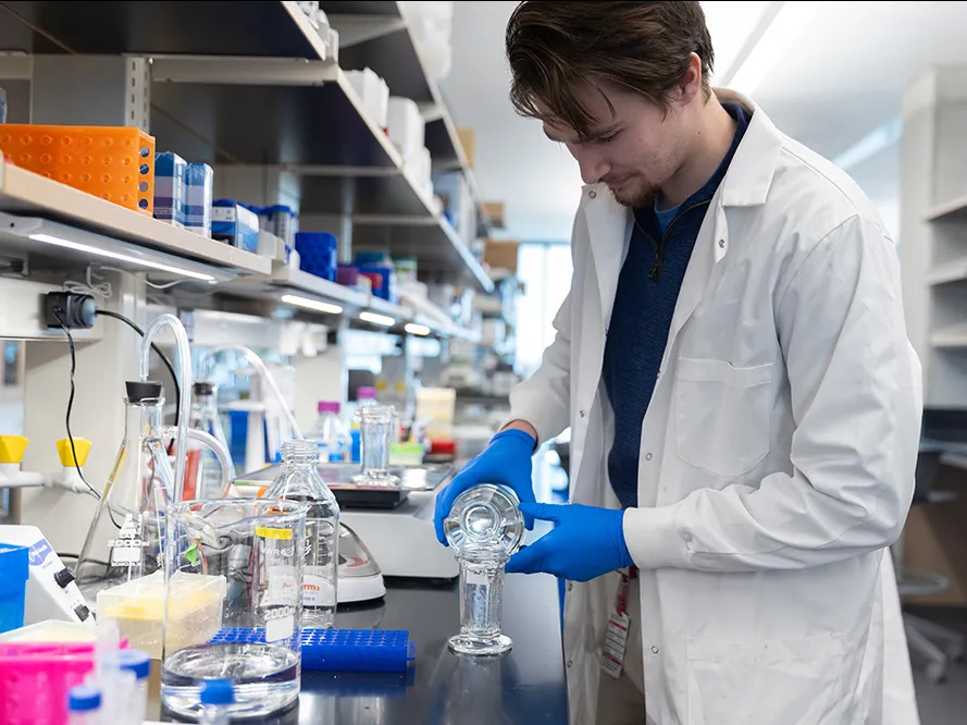 A white man in a lab coat works at a lab table with glass and colorful instruments and tools