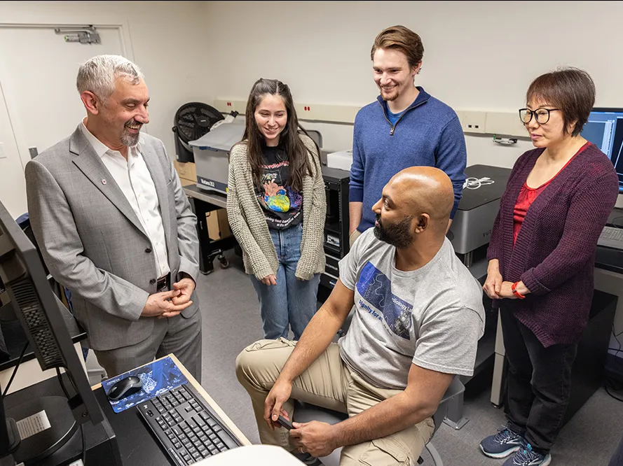 Five people of varying ages, racial origins and genders talk in front of a computer that shows images from a high-powered microscope. Those images can't be seen in this photo.