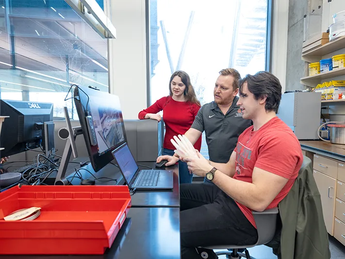 A student sits at a computer talking with others. From right to left, they go up a management level: the student, his boss (a white bearded man) and his boss, a young white woman.