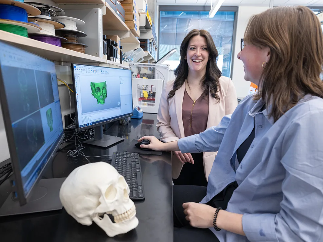 Two women talk in front on a computer monitor showing a 3-D skull. The 3-D printed result is on the table next to them.