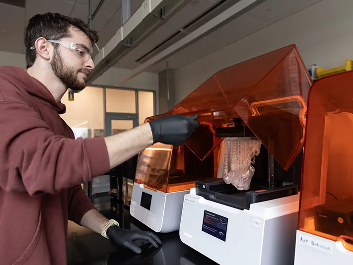 a young white man wearing a sweatshirt opens the transparent cover of a 3-D printer, which sits in a line of several. Each is about the size of a microwave stood on its side.. 