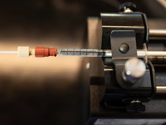 A heavy metal piece of equipment holds a slim tube filled with clear liquid. An even thinner line attaches to the end of the medicine tube