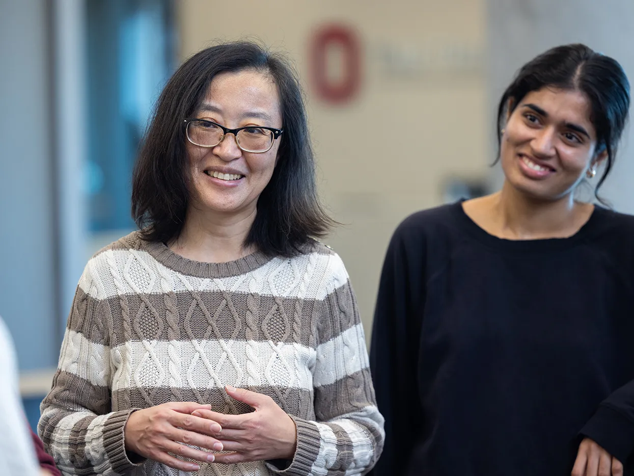 A woman of Chinese descent and slightly behind her, a woman of Indian descent, smile as they listen to an off-screen colleague speak
