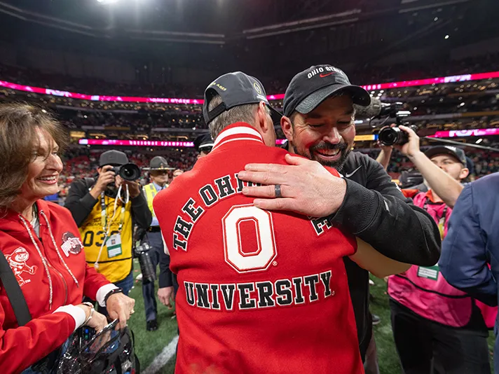 On a busy field after the game, football coach Ryan Day hugs the university's president, who can only be seen from behind. He wears a varsity athletics-style coat that says The Ohio State University.