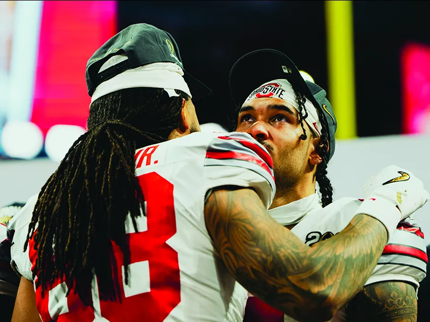 Two young Black men get ready to hug after their win. Both have long hair--the one whose back is to the camera has a bounty of braids halfway down his back pulled into a low ponytail. The player facing the camera looks overcome with emotion.