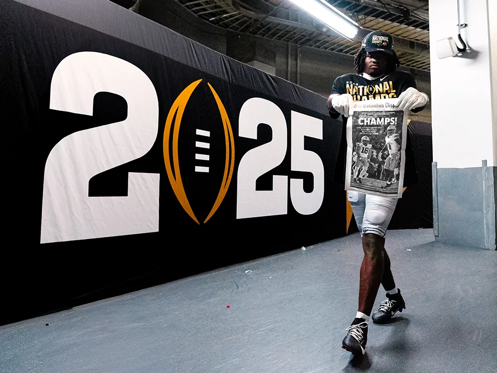 A young player holds up a traditional newspaper page that says Champs. He also wears a champs t-shirt over his football uniform, but his facial expression is mostly shadowed by his ballcap.