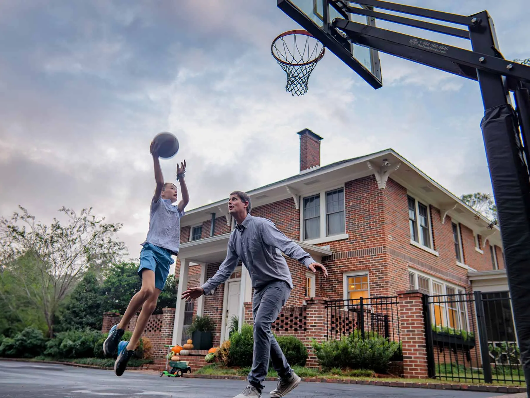 10-year-old Byron jumps as he shoots the basketball toward the net in a pickup game of basketball in the driveway at his home. He's a lanky white boy in gym clothes. His dad wears jeans and a button-up and they are intensely focused. Their brick two-story home is in the background.