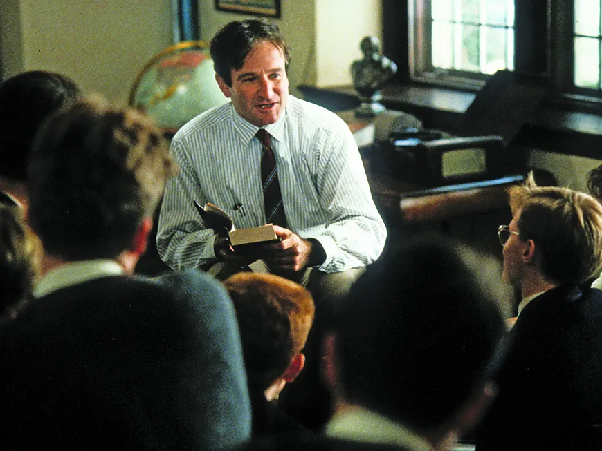 a teacher in a white button down and tie leans slightly toward his students, seen from behind, in this still from the movie Dead Poets Society.
