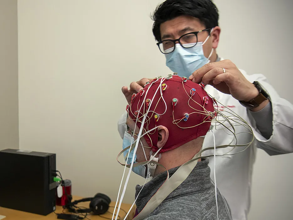 Dr. K. Luan Phan hooks up a stretchy cap with brain monitors to a patient reading a computer monitor. Dr. Phan is an Asian man with glasses and a lab coat. The patient is an older white man. Both wear paper masks.