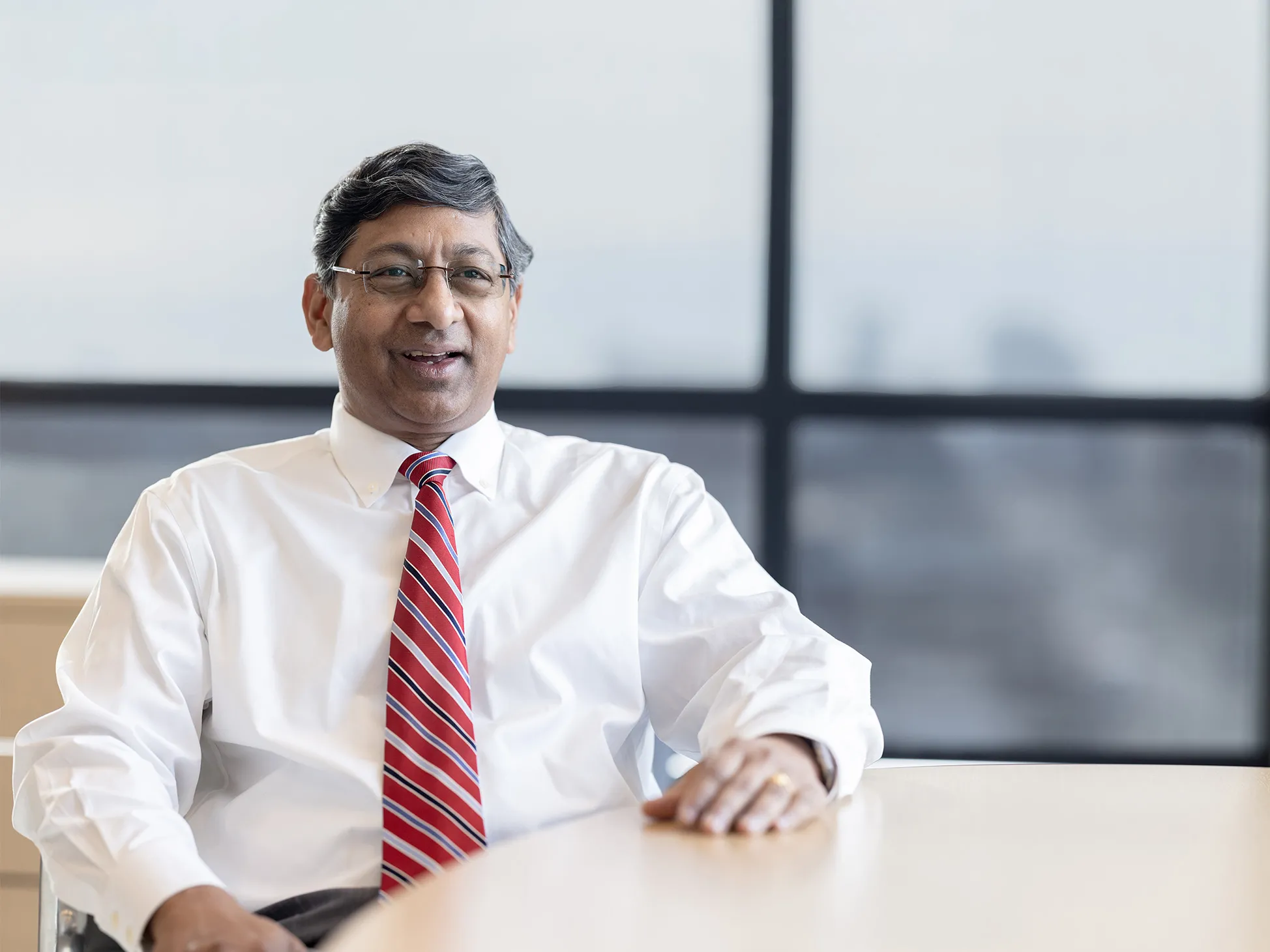 Ravi Bellamkonda, a middle-aged man of Indian descent, sits casually at a table in front of windows overlooking campus. He smiles as he talks.