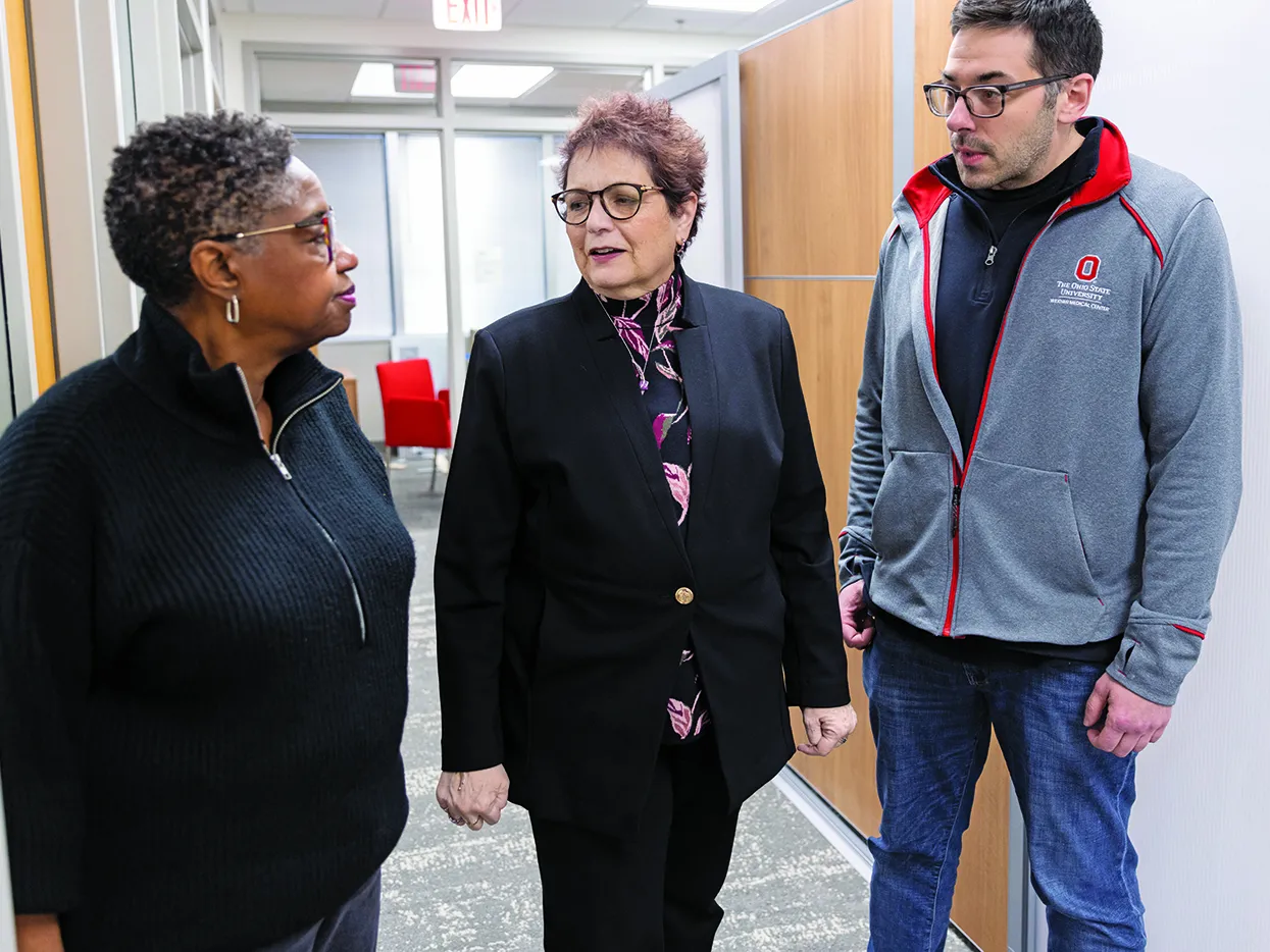 Pausing in an Ohio State building hallway, Electra Paskett speaks with two colleagues. She's a white woman with short hair, glasses and a stylish suit; to her right is a Black woman with short hair and to Electra's left is a tall white man in an Ohio State fleece sweatshirt. They seem invested in their conversation.