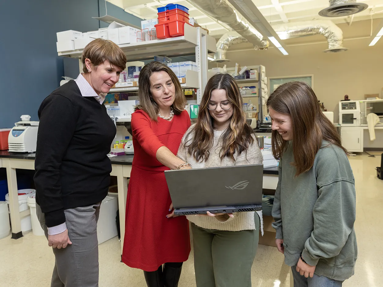 Dr. Tamar Gur, wearing a red dress, points at a laptop computer screen held by a young woman with long hair. Two more people are in the photo looking at the computer. They're all smiling and appear as though they enjoy working together.