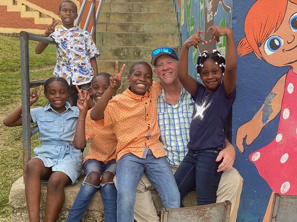 A man sits with five kids on stairs next to colorfully painted walls. They're all grinning and the kids are making peace or heart signs.