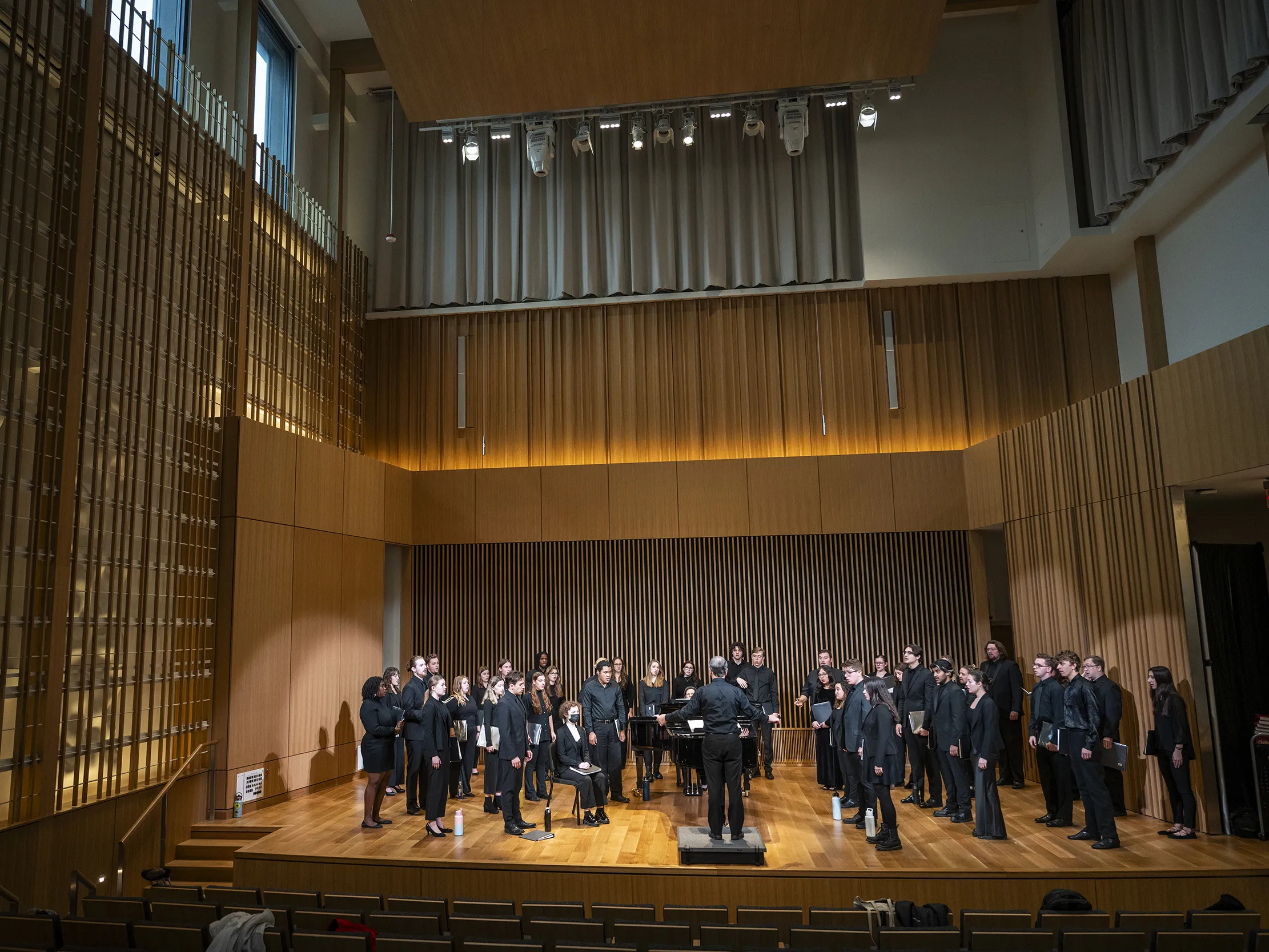 Students standing in rows wearing all black performance outfits singe on a wooden stage as a conductor holds his hands wide. The stage is wrapped in sound optimization and modern-looking elements. The lighting is very warm.