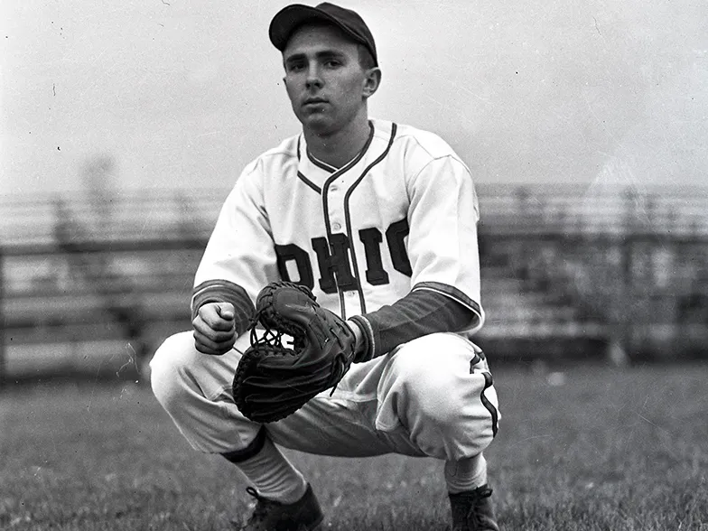 In a historical photo, a baseball player crouches and wears a serious expression as he is photographed. The front of his jersey says Ohio and his mitt is thick with an extra large thumb portion.