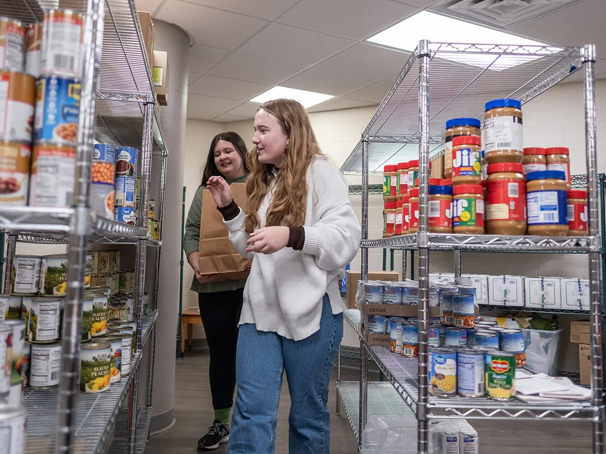 Two student workers, young white women in casual clothes, choose goods from shelves at the student food pantry.