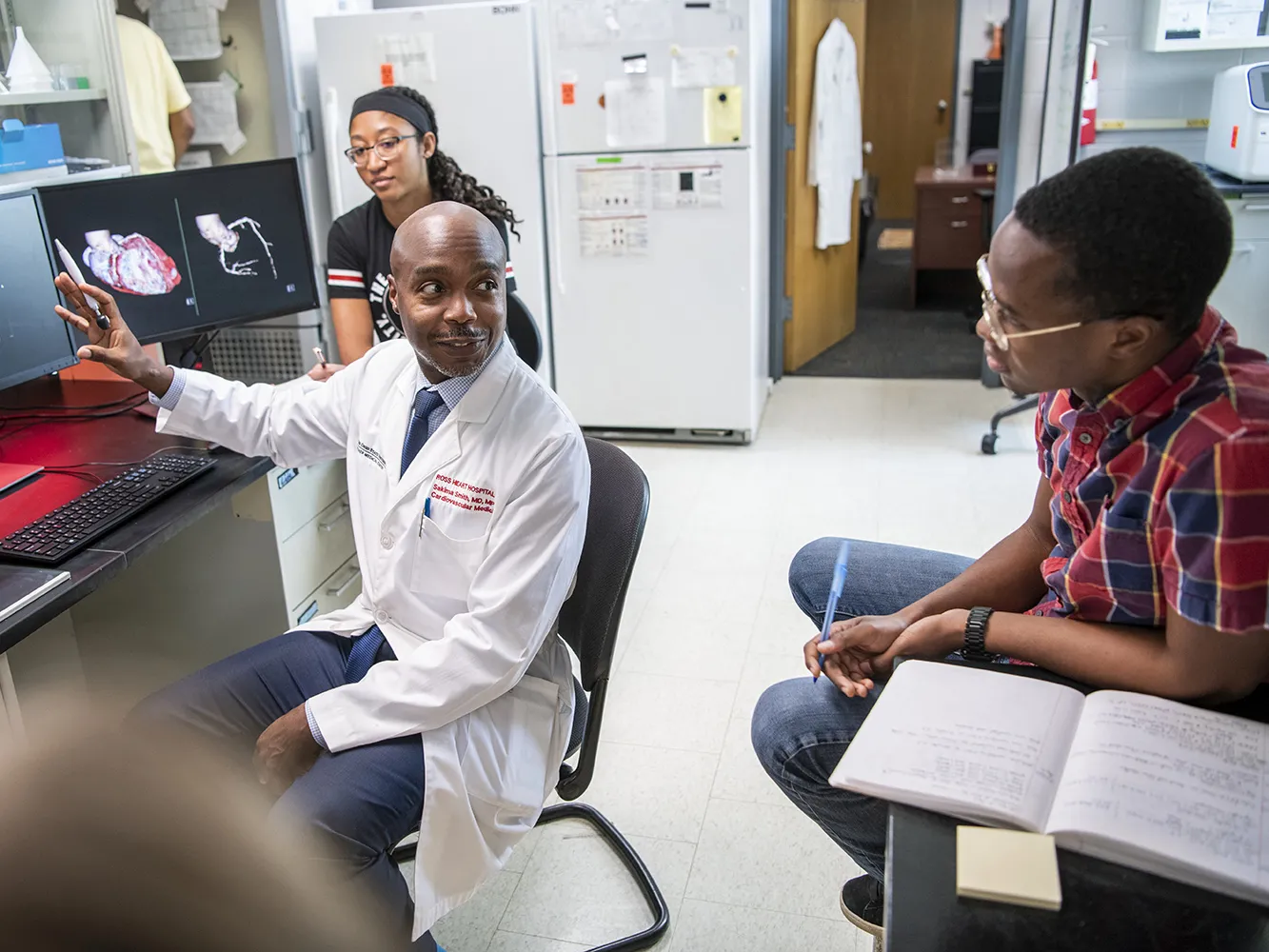 Dr. Sakima Smith, a distinguished looking black man, gestures as his monitor as he teaches a small group of students in a laboratory setting.