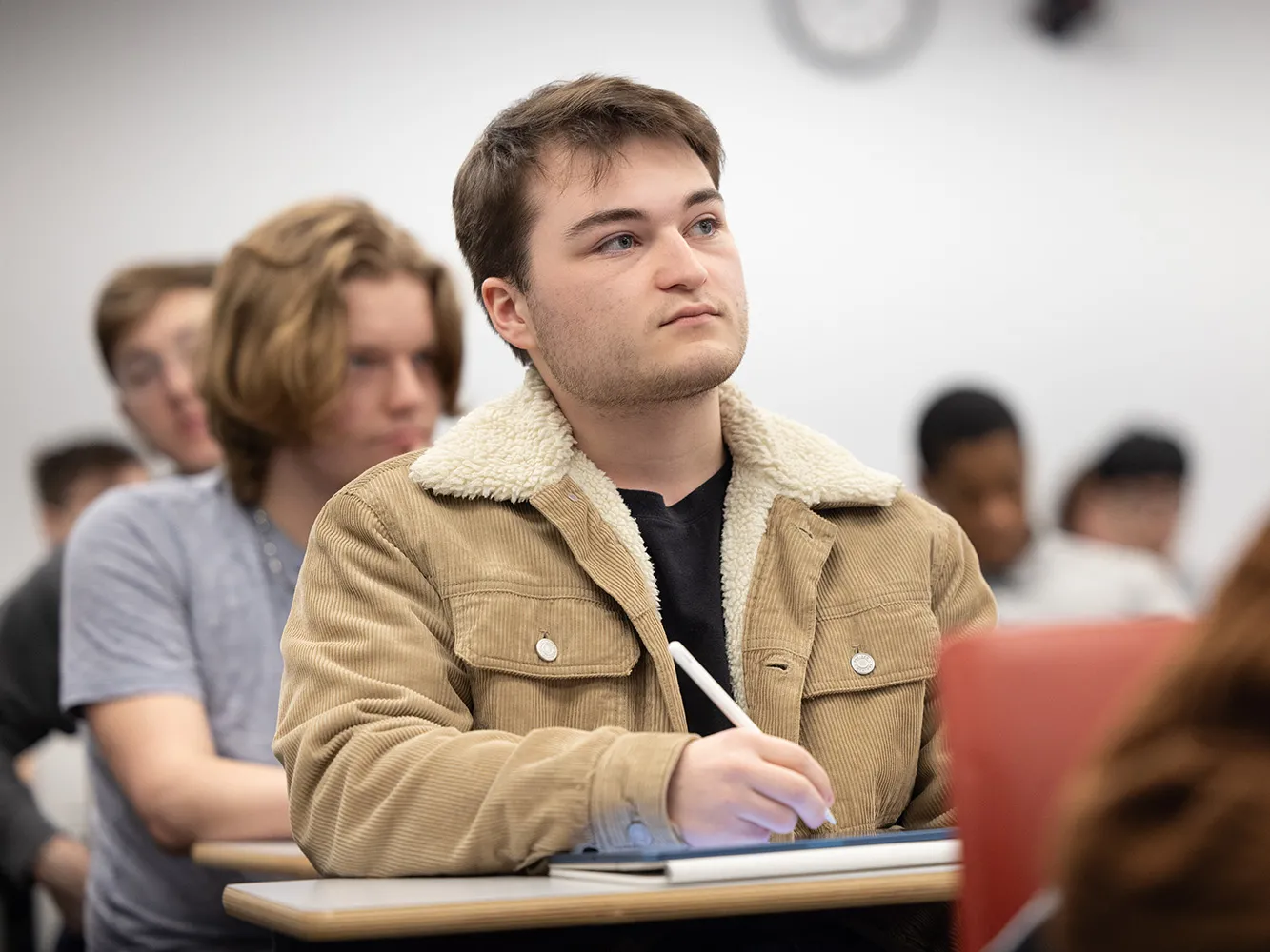A young white man sitting in a filled college classroom intently listens to his professor (who's not in the photo) and takes notes