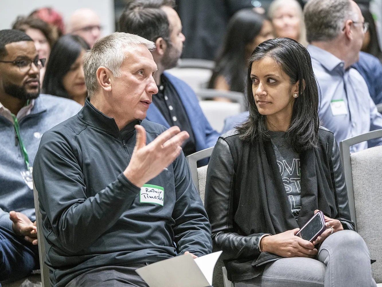 Ratmir Timashev, a white man with short white hair, gestures as he sits at an event and speaks to the woman beside him, Shereen Agrawal. She is a petite woman on Indian descent wearing a chic jacket over her T-shirt and Jeans. They're discussion seems serious, and many people of all races and genders fill seats behind their row.