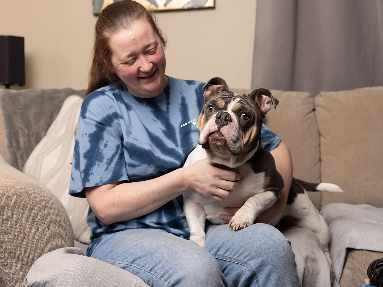 A woman smiles as she looks down at the dog she's holding on her lap. Her pet is an English bulldog with charming face wrinkles and multicolored spots. It is slightly bug eyed. All together it looks like it'd enjoy some pets from the viewer.