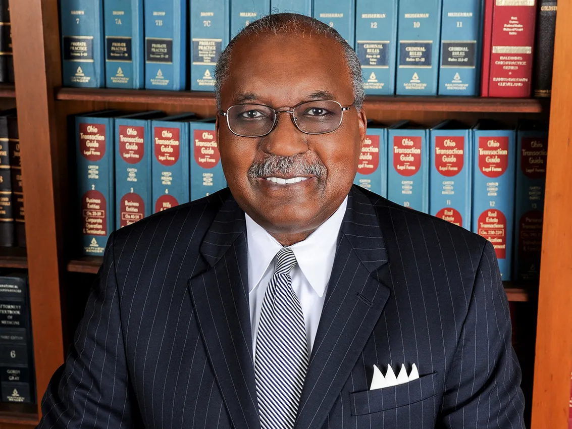 This portrait of Ben Espy shows a sophisticated Black man with a friendly smile. He wears glasses and a pin-striped suit and stands in front of shelves lined with law books.