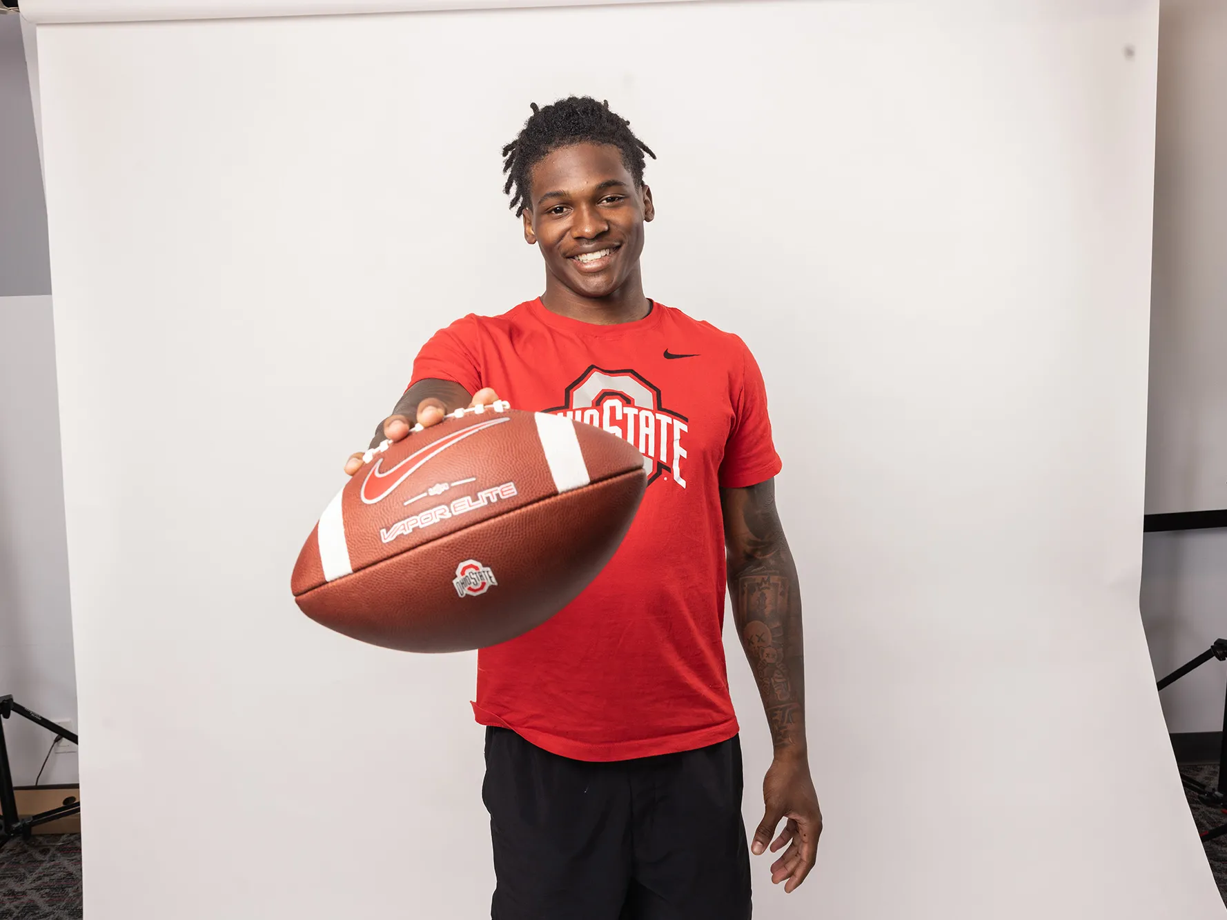 Ohio State football player Jeremiah Smith poses for a photo holding out a football. He's an attractive young black man, smiling with his hair pulled up in a ponytail and tattoos on his arms.