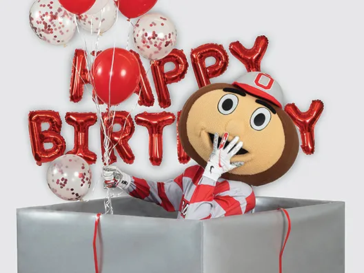 Brutus Buckeye puts a hand to his mouth as he giggles while rising from inside a giant gift-wrapped box.