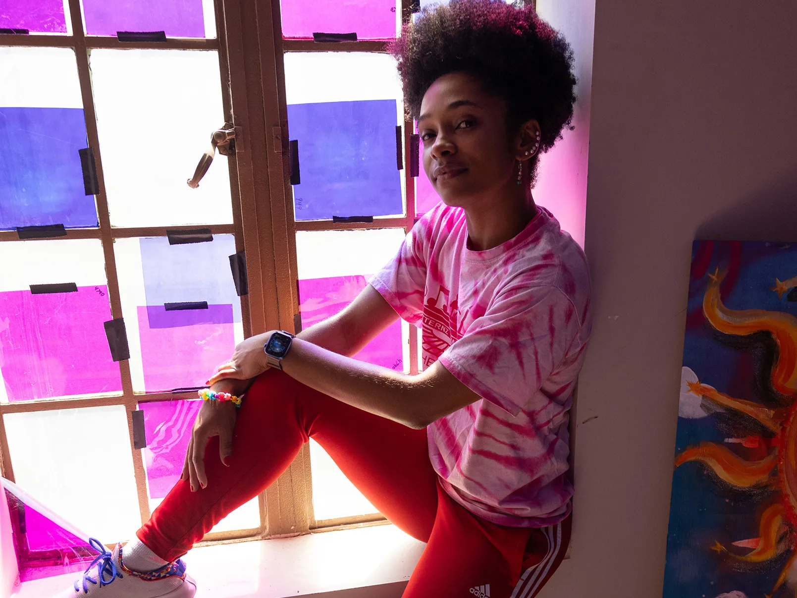 Brianna Rhodes, a pretty, young black woman, sits on a window ledge inside a dim room. Colored paper taped over the window shines colored light onto her. She's smiling with her eyes and wearing casual clothes that she can dance in. Her hair is tight curls on top of her head and she has a line of earrings up the ear the photo shows. 