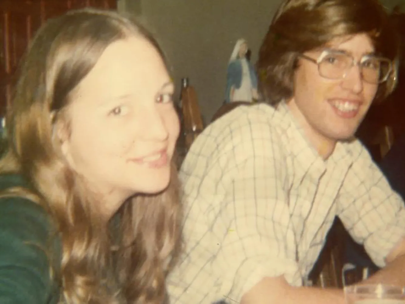 In this 1970s photo, a young white couple smiles at the person taking the picture, almost as if they're saying "if you really have to take this photo, go ahead." He wears glasses and she has long hair--their hair color almost matches.