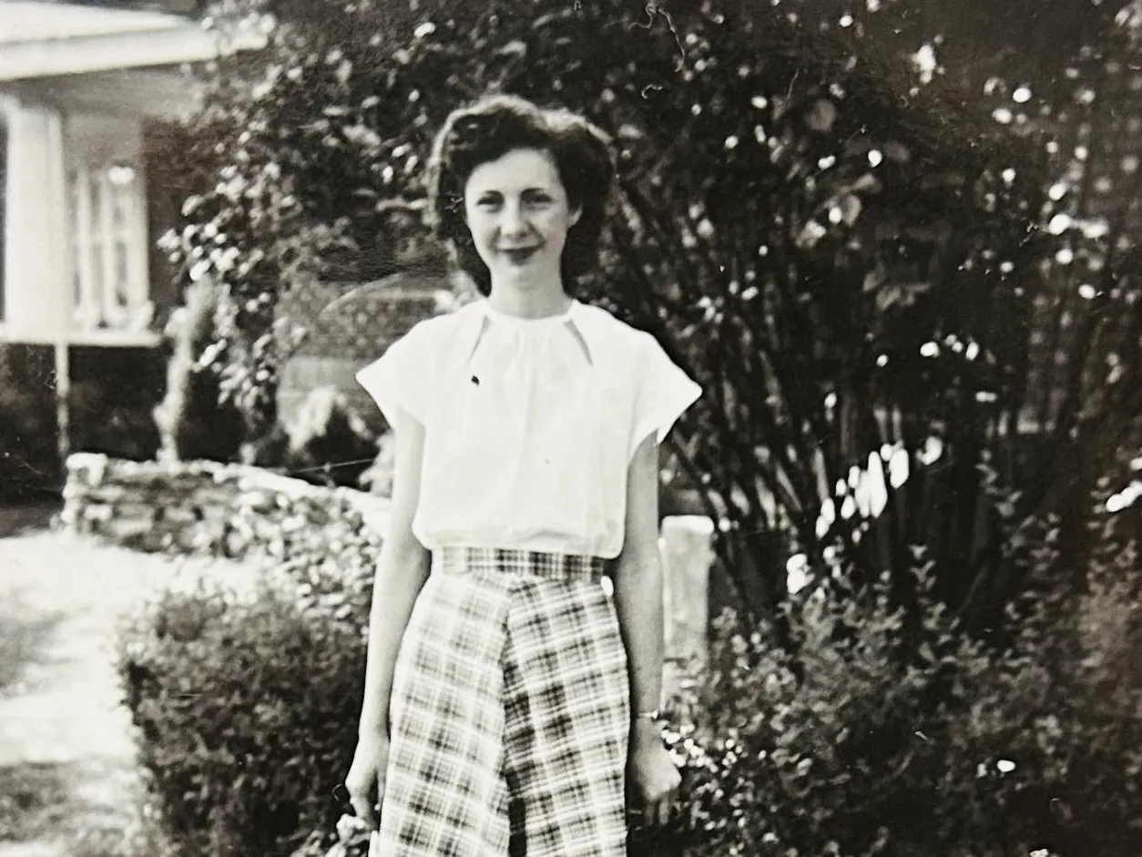 As a young woman in this black and white photo, Ruth Klingler McPheron stands in front of a house and greenery wearing a white blouse and shin-length plaid skirt. She is a white woman with a pretty hairstyle, a soft smile and the looks of someone you'd want to tell stories to.