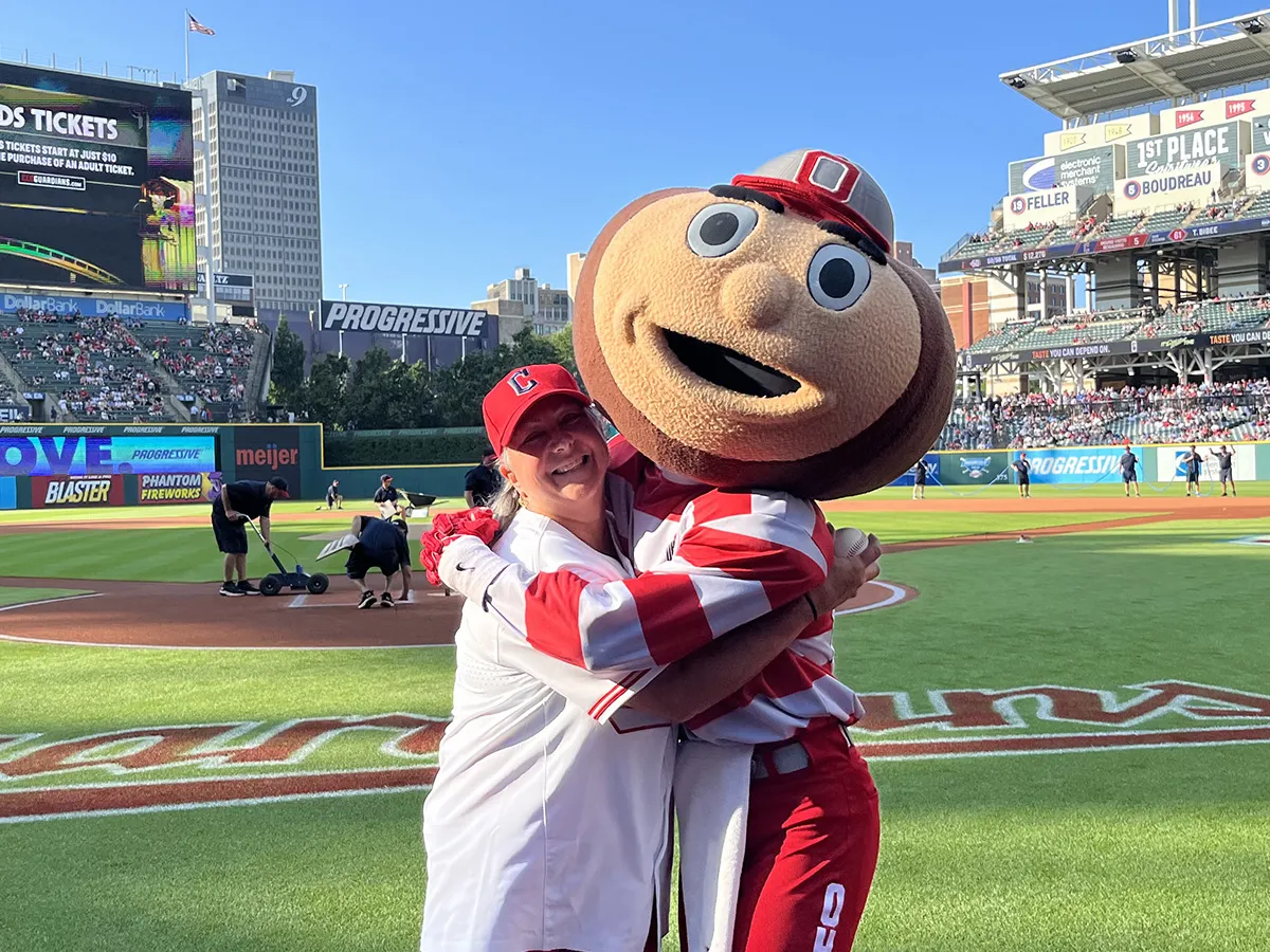 Molly Ranz Calhoun, wearing a baseball cap, T-shirt and shorts, laughs as she gives Brutus Buckeye, Ohio State's mascot with a buckeye-shaped giant head, a hug at the Cleveland Guardians' baseball stadium. Molly is a middle-age white woman with shoulder-length hair and a friendly smile.