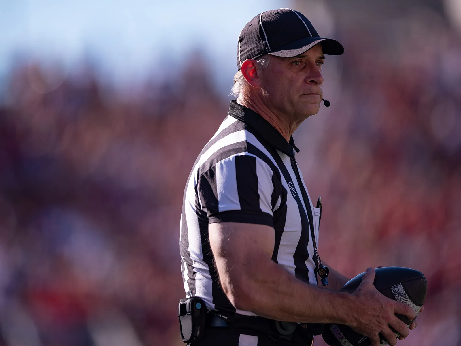 A football referee holds the ball and makes a thoughtful face as he watches something out of the photo frame. He wears a black and white striped referee’s shirt, a ballcap and a headset that leaves a small mic in front of his mouth to communicate calls. He's a white man with white hair and muscular arms. 