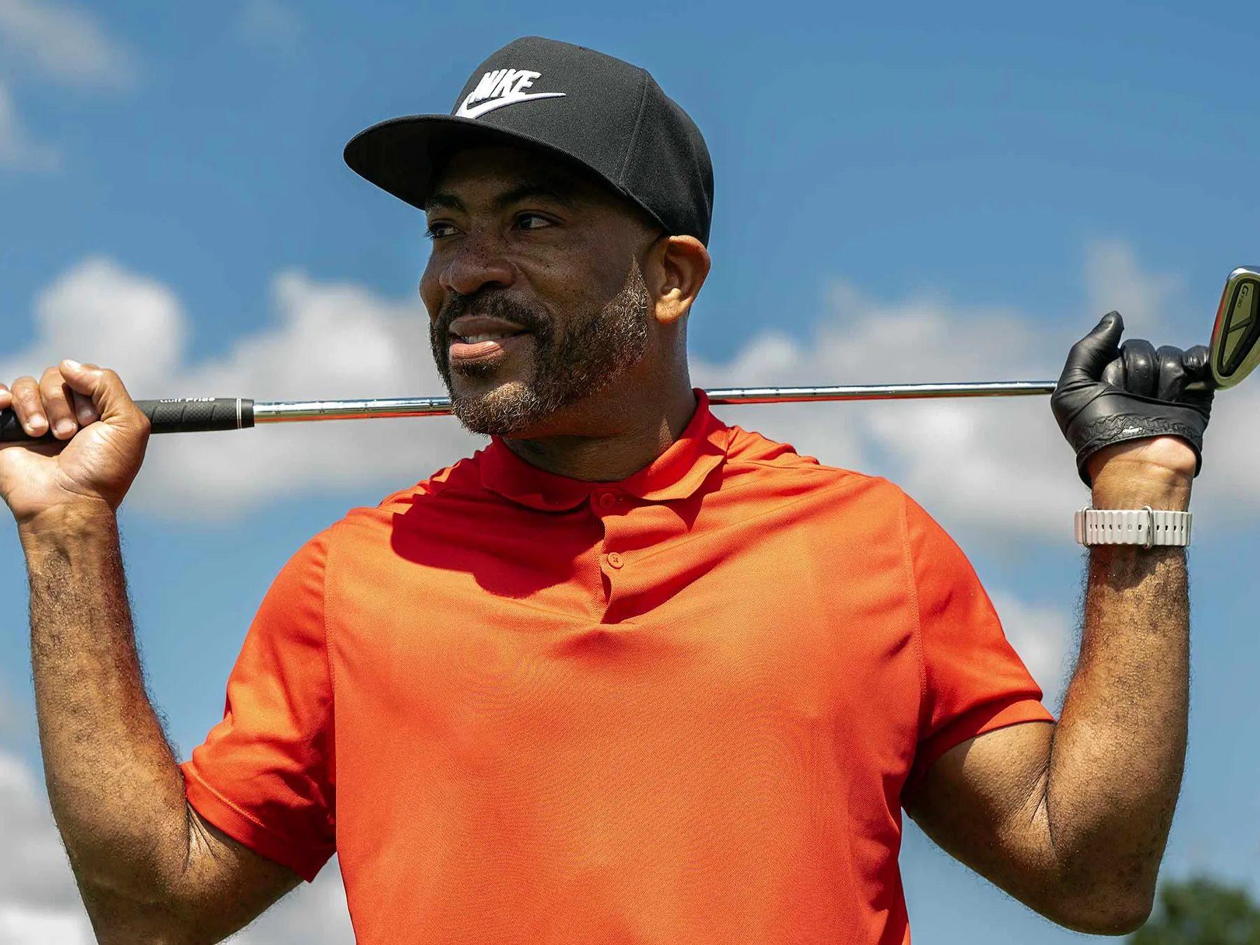 Ona golf course, Dee Miller smiles as he holds golf club behind his head. He wear a Nike baseball cap and a bright golf shirt.