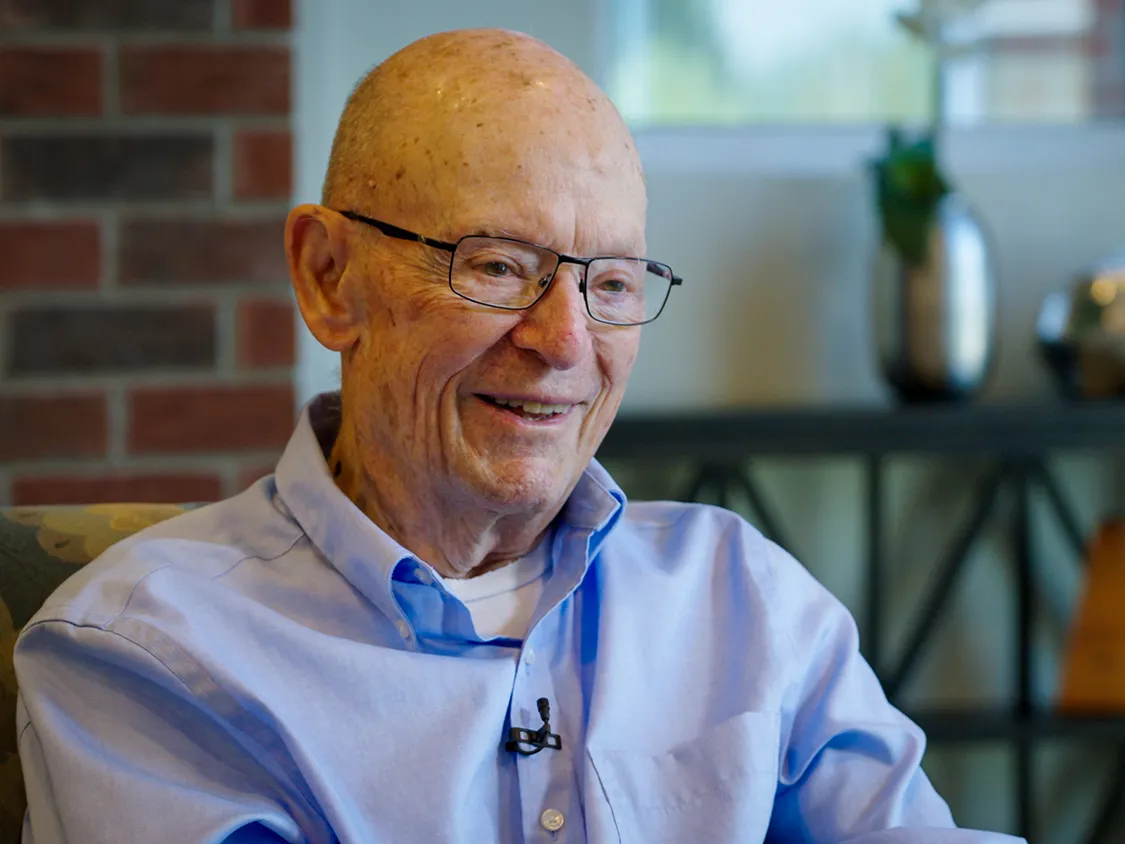 Dick Wood smiles as he tells a story. He's an older white man wearing glasses and a button-up shirt. Lines on his face shows he's smiled a lot through his years.
