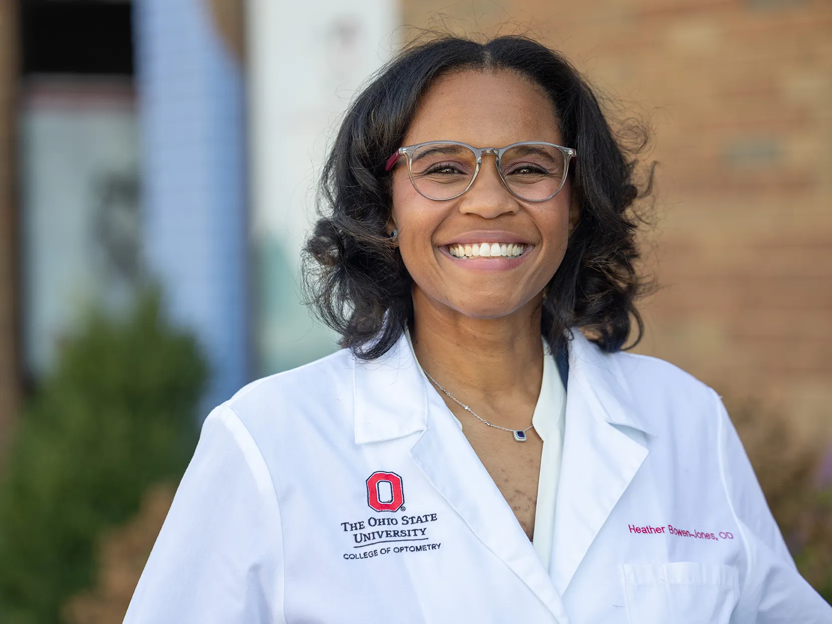 Heather Bowen-Jones ’11 OD grins for the photographer as she is photographed for a portrait. She stands in front of a medical building, wears glasses and a white doctor's coat, and she looks genuinely happy to share what she does.