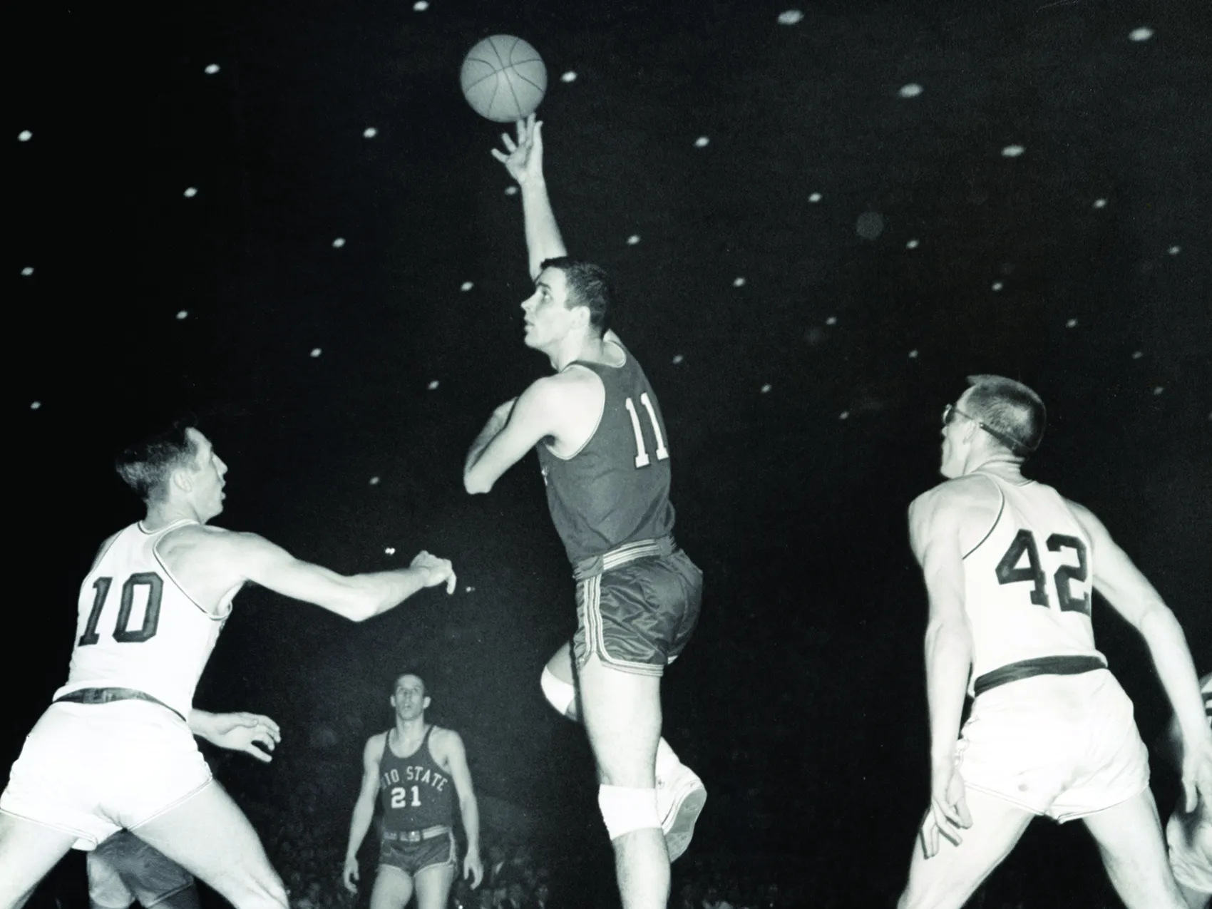 In an older black and white photo, an Ohio STate basketball olayer (Jerry Lucas) jumps high off the floor of the basketball court and hooks the ball over his head. Players from other teams look mini as he rises so high above them. His jersey is number 11 and he's. ayoung white man with neatly shorn hair and a strong build. He also wears knee pads.