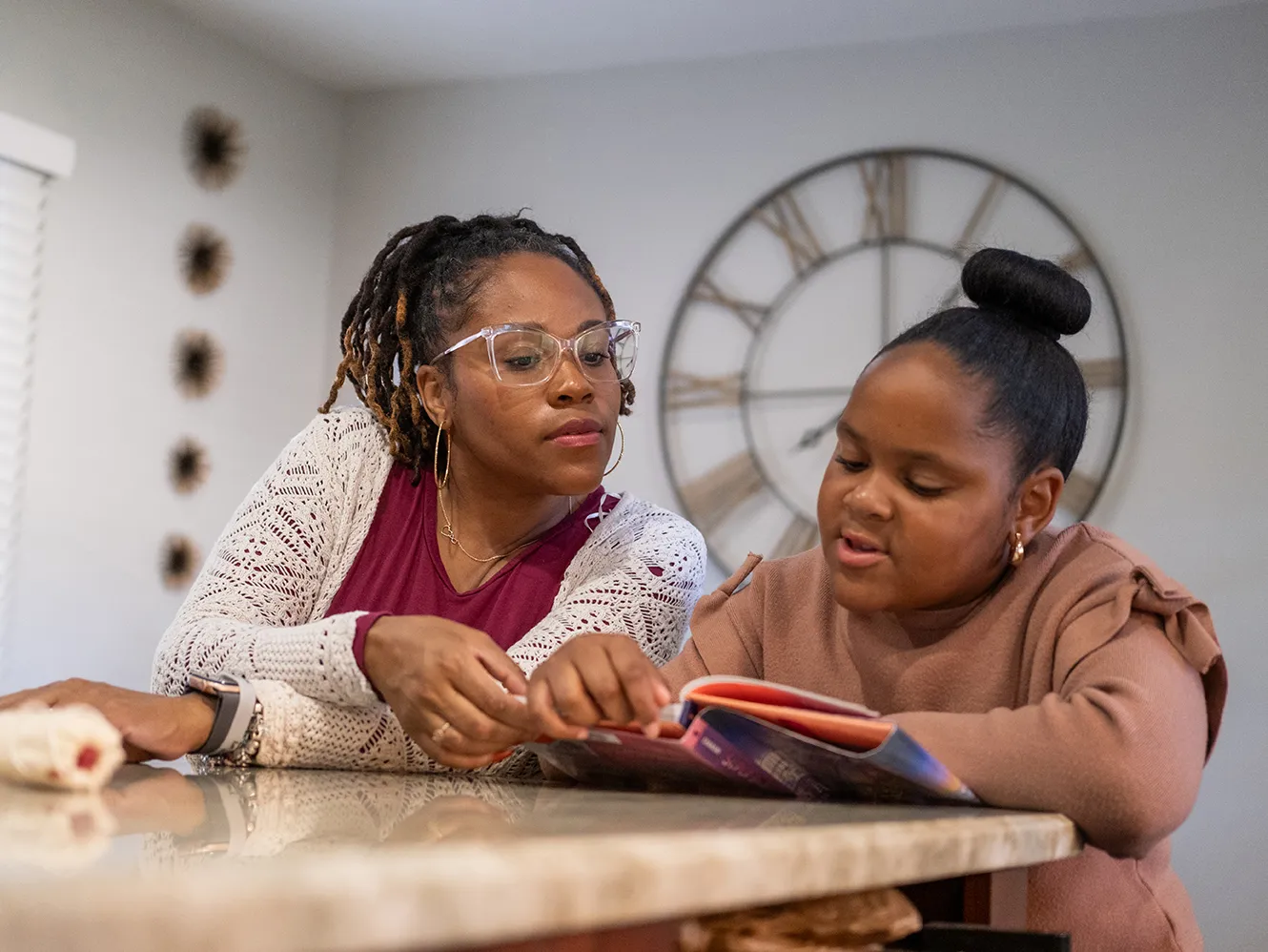 Nine-year-old Harper reads a thick children's book as her mom leans in to see the page. Harper is a cute Black girl with her hair pulled into a neat bun on top of her head, and her mom wears glasses and a pretty cardigan. They sit in their kitchen at home.