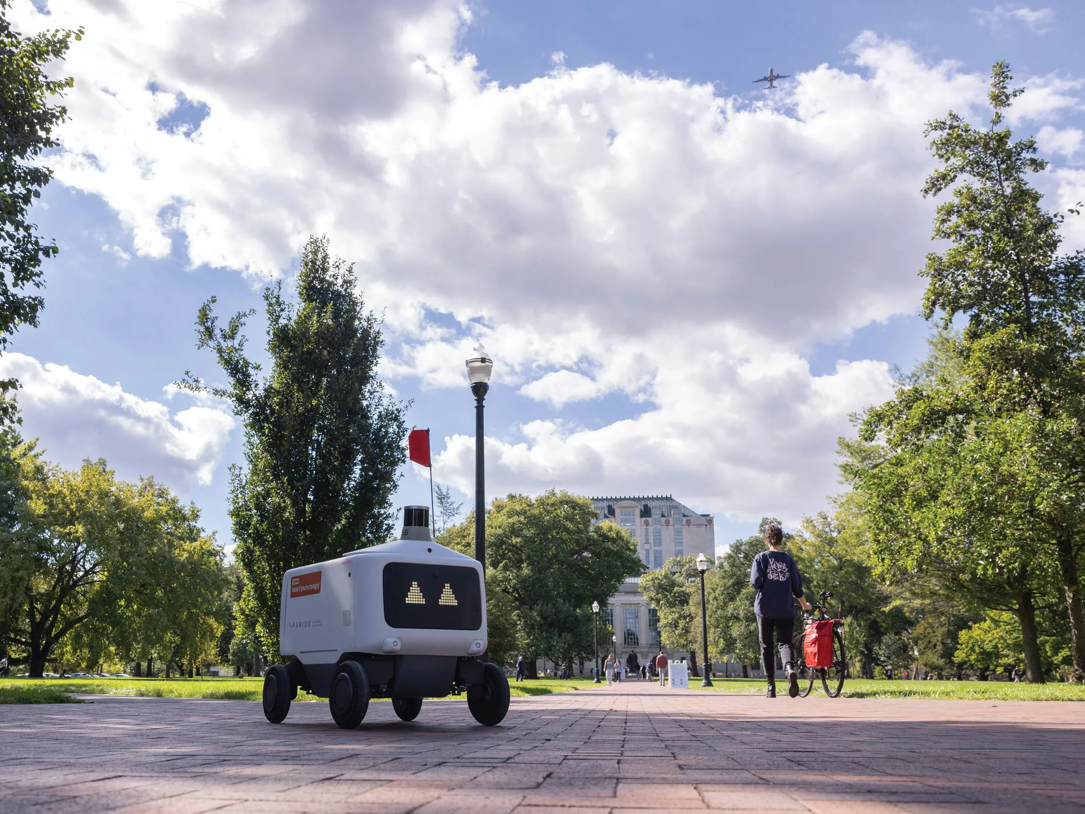 A lone food rover, which is a plastic-covered rectangle with a screen face showing triangle-shape eyes, makes its way across the oval. Thompson Library and students can be seen in the distance.