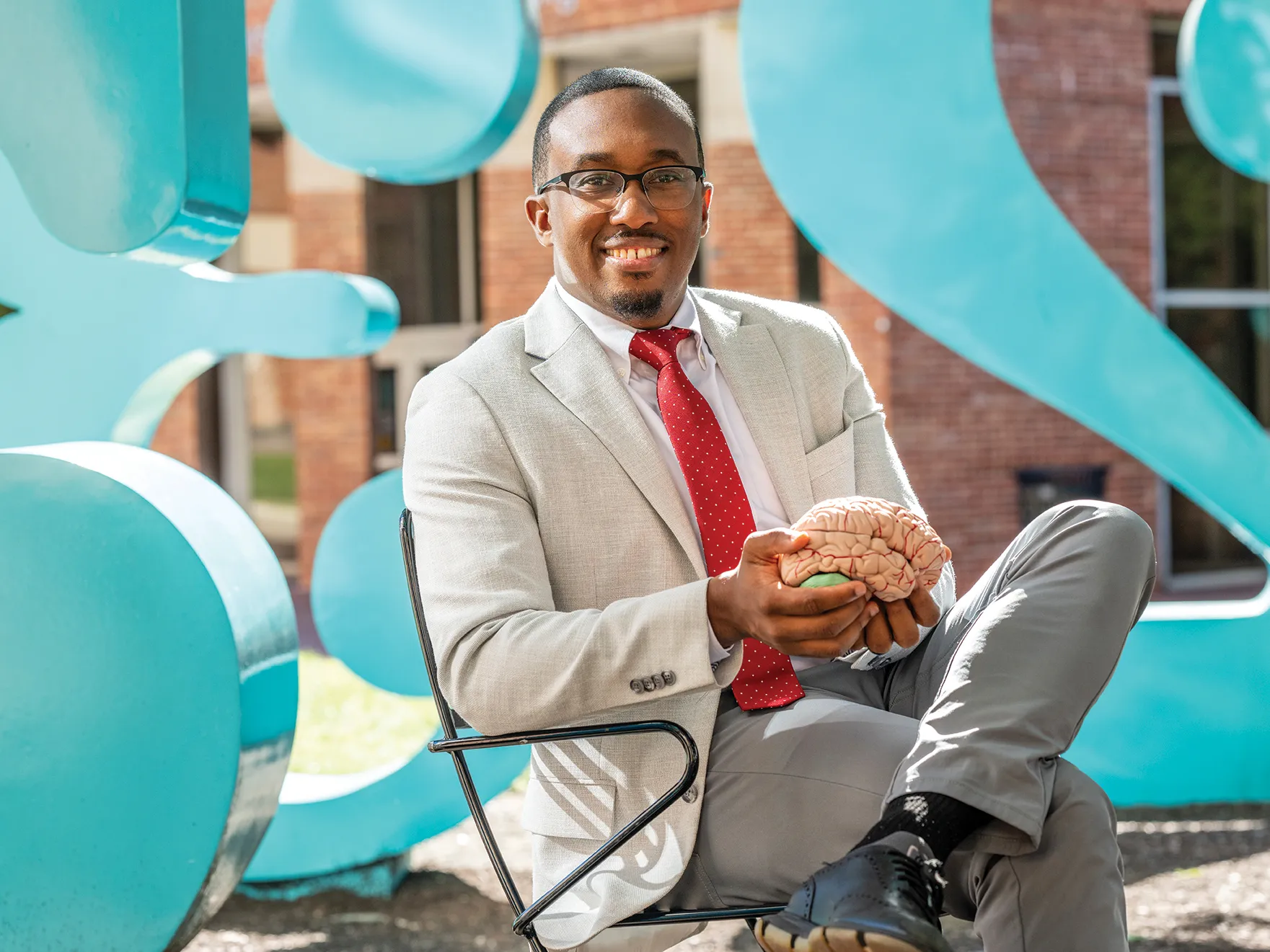 Jeremy Grant holds a model brain while sitting in a chair at the numbers sculptures on Ohio State's campus. He's a black man wearing a suit, tie and glasses. He has a friendly smile and his hair is short and neatly trimmed. 
