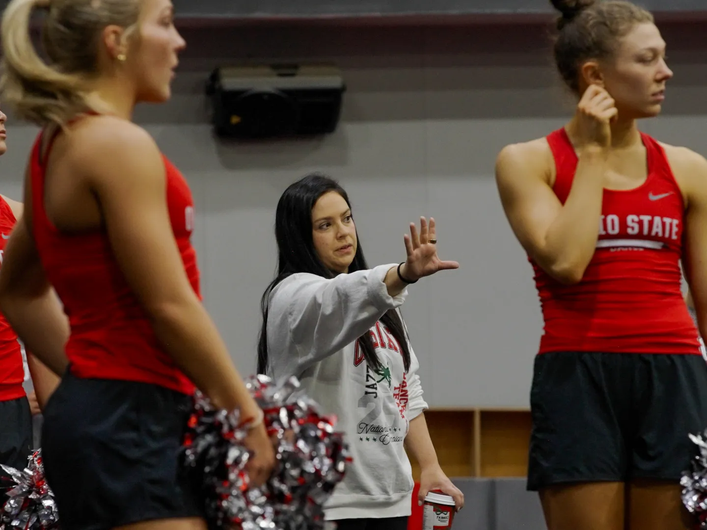 Melissa McGhee, a short, pretty white woman with long dark hair, coaches dance team members. She gestures with her hand reached out and they all look that way.