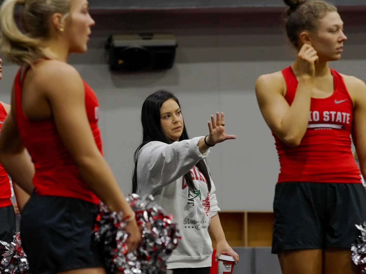 Melissa McGhee, a short, pretty white woman with long dark hair, coaches dance team members. She gestures with her hand reached out and they all look that way.