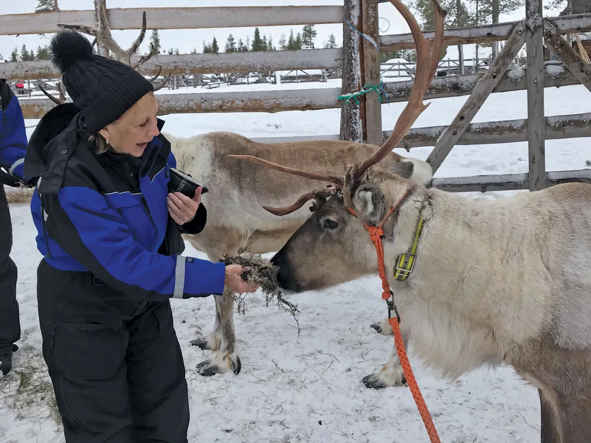 A white woman in snow gear feeds a reindeer on a trip. They're surrounded by snow.