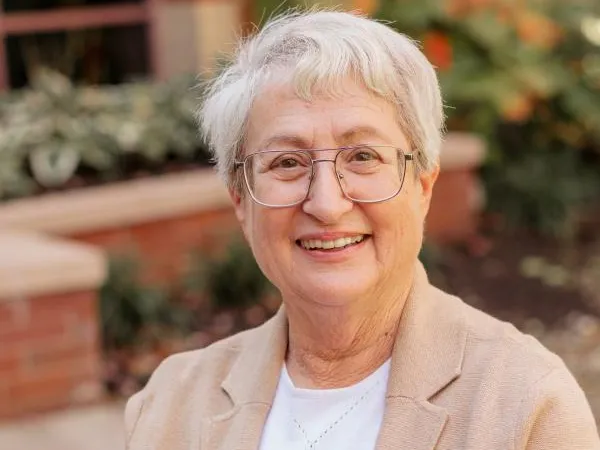 A white woman with short gray hair, glasses and a friendly smile poses for this photo wearing a T-shirt and suit jacket.