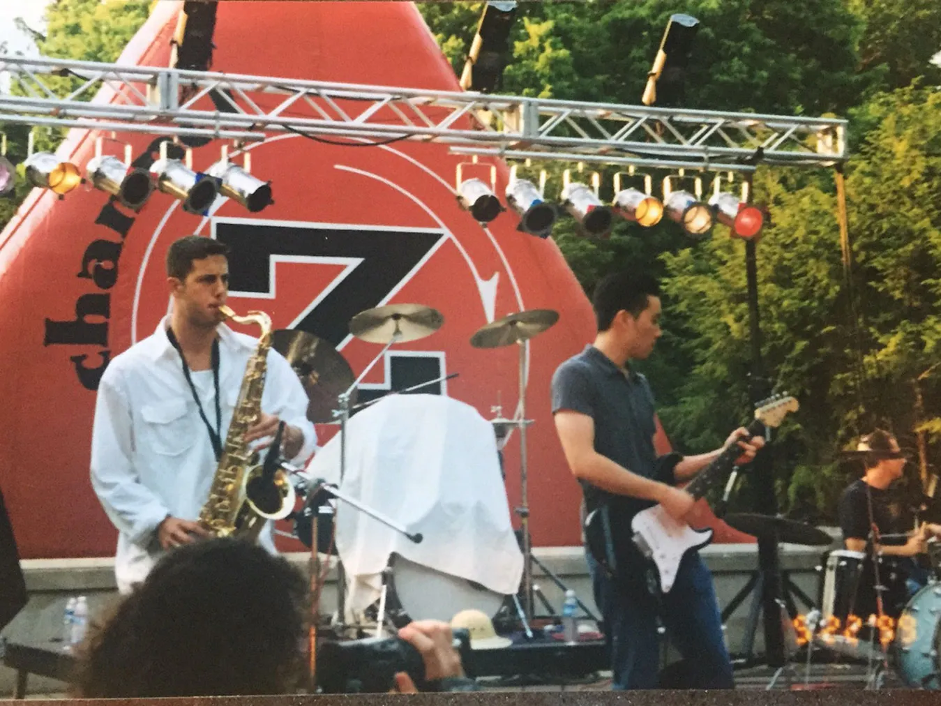 At a small Columbus festival, several musicians perform on an outdoor stage. One plays a saxophone, another plays an electric guitar, and a third plays a drum set. Trees fill the background.