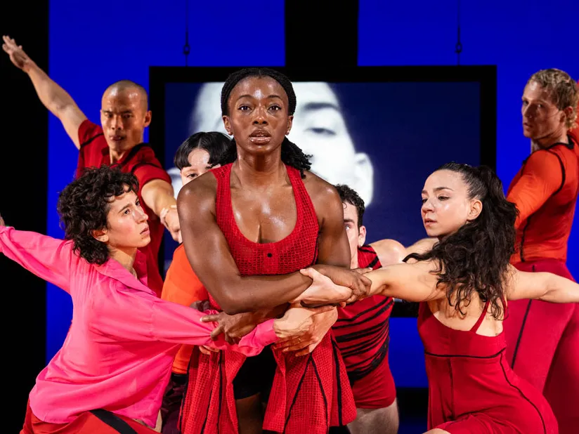 A group of dancers onstage perform a synchronized pose, holding onto one another in an interconnected formation. They wear brightly colored costumes. Behind them, large screens display black and white images, adding contrast to the vivid lighting and dynamic movement onstage.