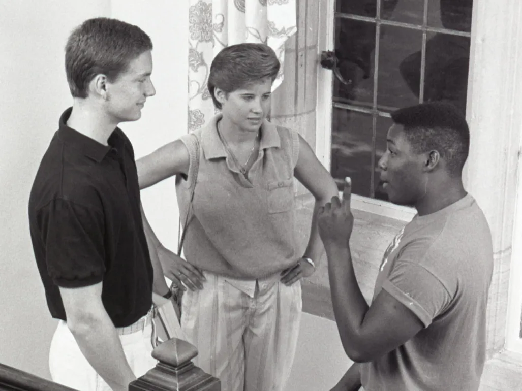 On a staircase landing, three college students engage in conversation. A large window with patterned curtains is behind them, and the wooden staircase features turned balusters and a decorative newel post. Of the three, one is woman and two are men; one is Black and two are white. They’re dressed in styles common in the 1980s.
