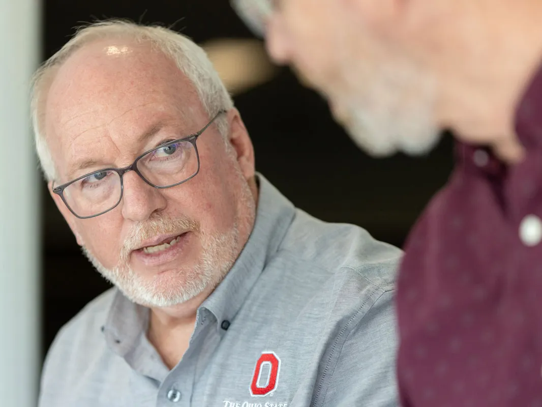 Dale Gelter talks to a colleague with a serious expression. He’s an older man with neat white hair and glasses and wears a button-down shirt with an embroidered Block O.