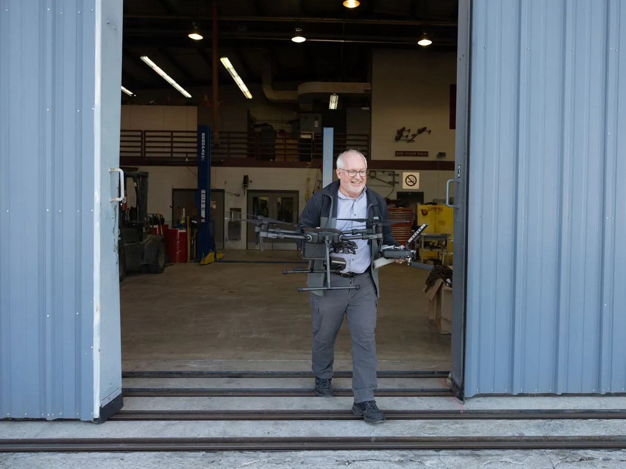 Dale walks out of the shop doorway while carrying a multi rotor drone with both hands. The scene is framed by the open metal doors of the structure.