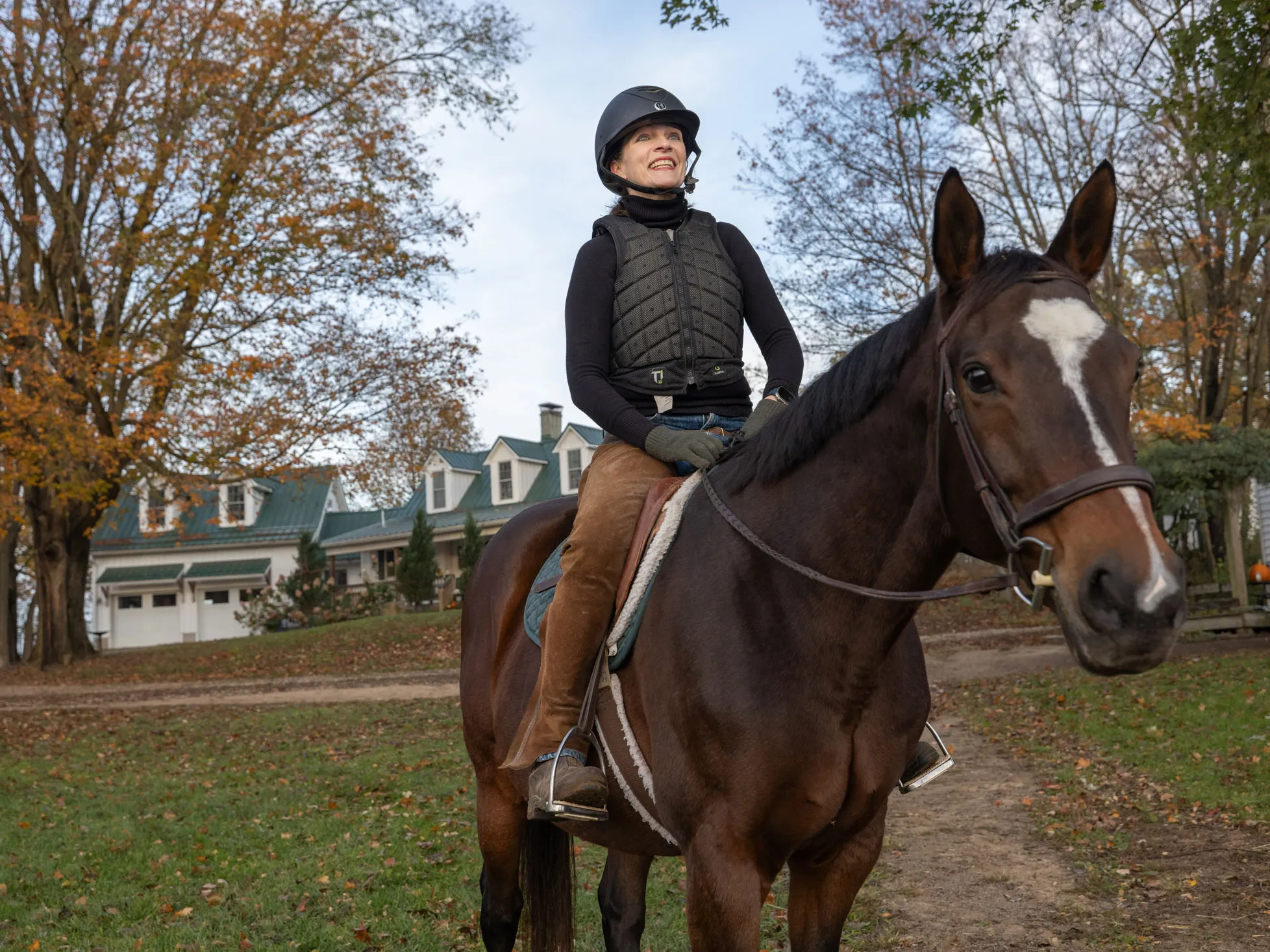 Kathy Lloyd smiles as she sits on a horse outside her riding stables on a crisp autumn day. She wears a helmet and protective riding vest sits and the horse looks directly at the camera as she looks off into the distance.